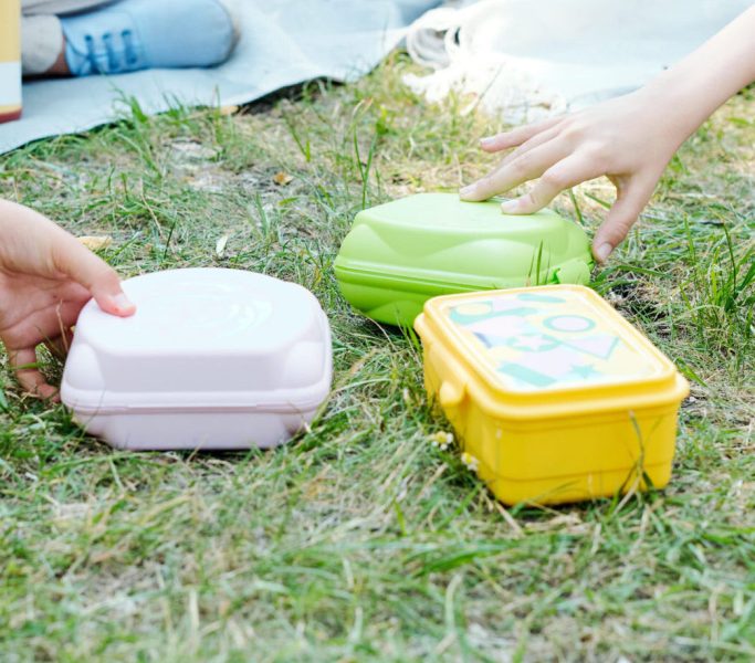 Close-up of unrecognizable children taking lunch boxes from grass while having picnic together outdoors